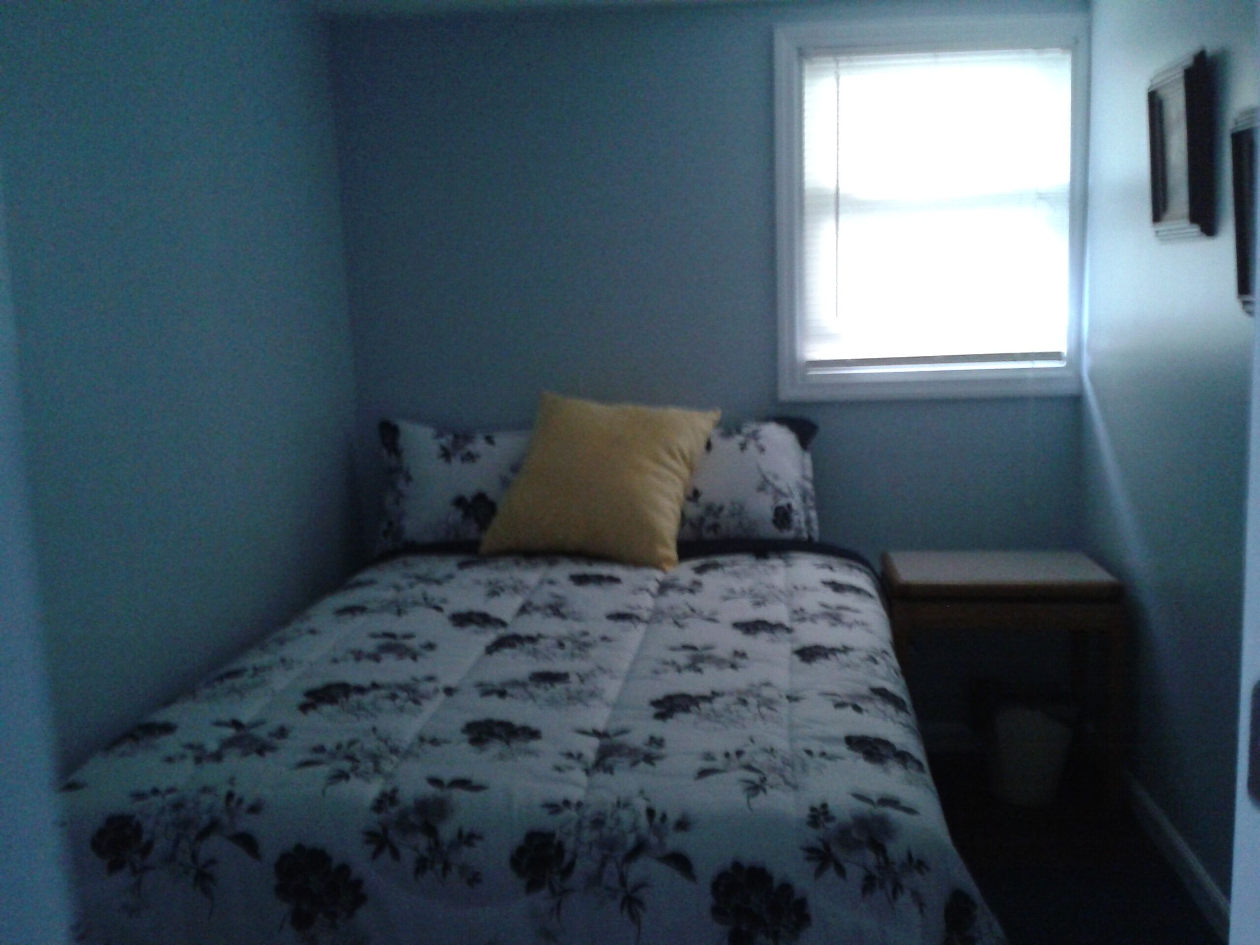 A small, single-bed room with a floral bedspread, a yellow pillow, and a window providing natural light.