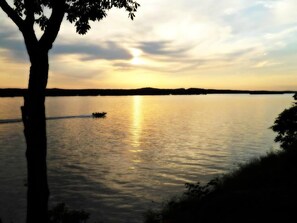 Ein ruhiger See bei Sonnenuntergang, im Hintergrund ein Boot und im Vordergrund ein Baum.