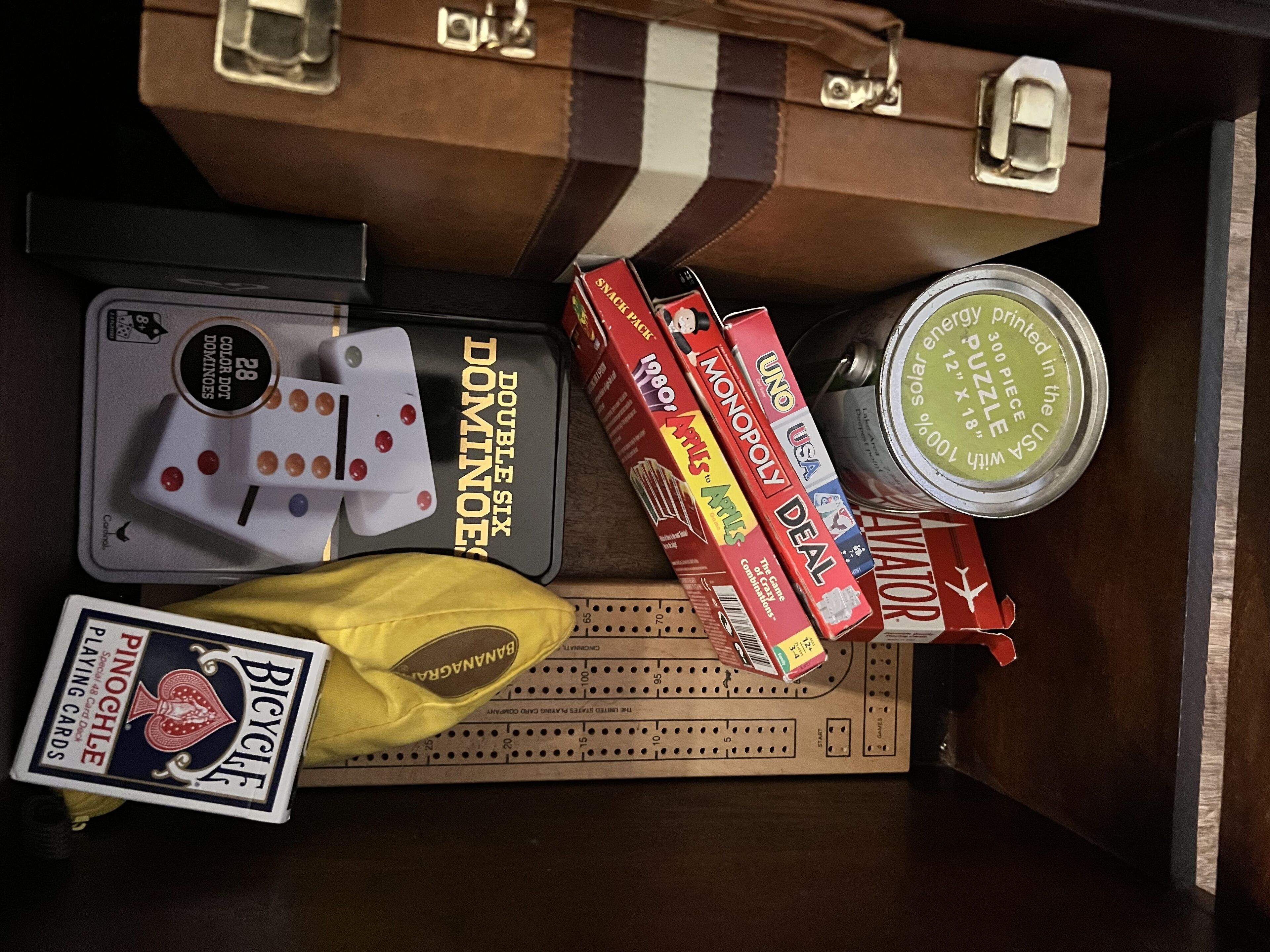 Uma mala com padrão listrado marrom e preto, um jogo de Banco Imobiliário, um conjunto de dominós, um baralho de cartas e uma caixa de quebra-cabeça.