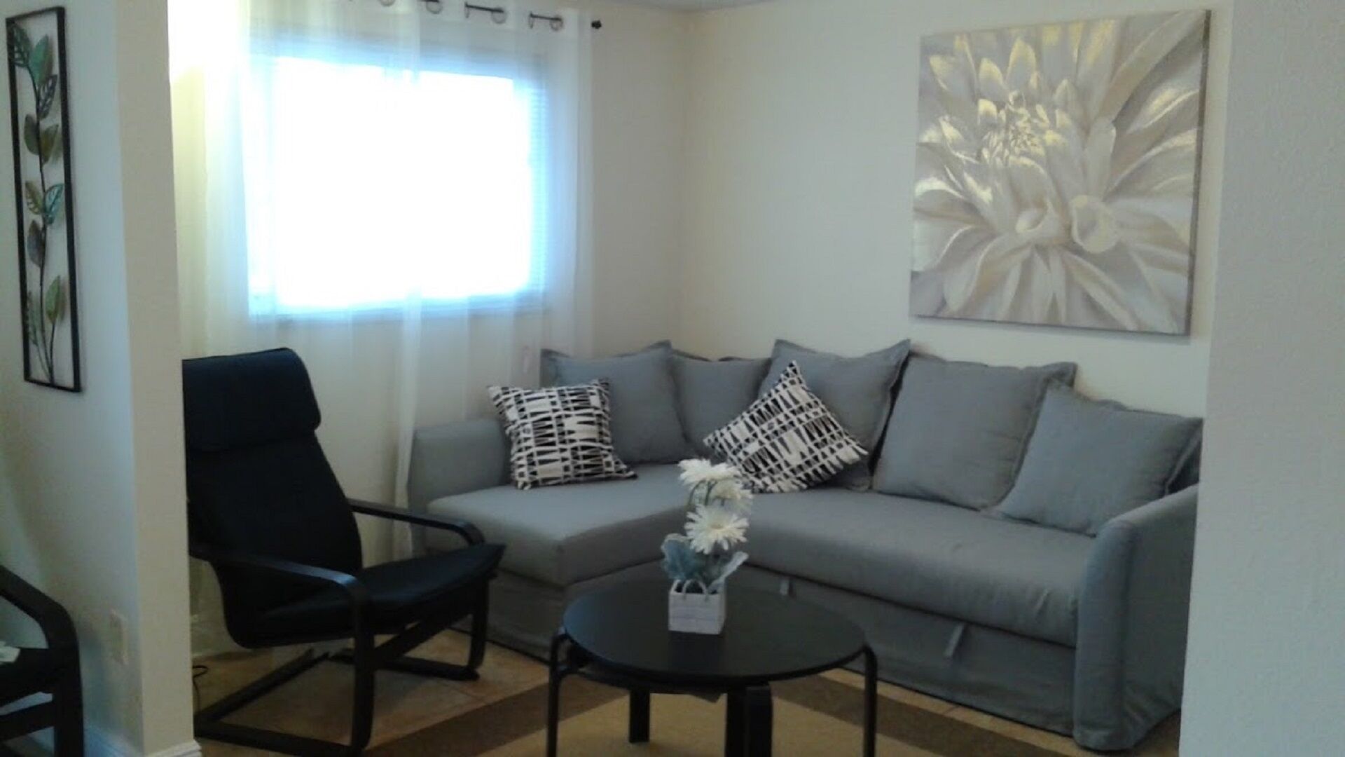 Ein Wohnzimmer mit grauem Sofa, Kissen mit schwarz-weißem Muster, einem schwarzen Sessel und einem kleinen runden Tisch mit einer Vase.