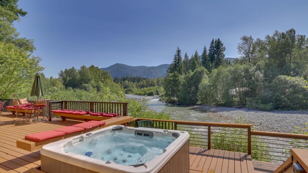 Une terrasse dotée d’un jacuzzi, de sièges rembourrés de couleur rouge et offrant une vue sur une rivière et des montagnes boisées.