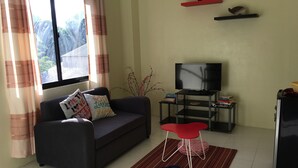 A living room with a sofa, a TV on a stand, a small table with books, and a window with curtains. - Cozy private apartment - Walk to the center (Dumaguete City)