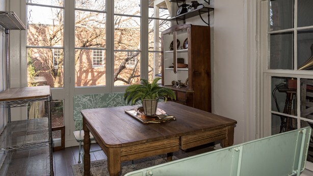 A dining area with a wooden table, a potted plant, and a view of a brick building through large windows. - Family-Friendly 1830s Cville Row House Near Downtown Mall, UVA, & Monticello (Charlottesville)