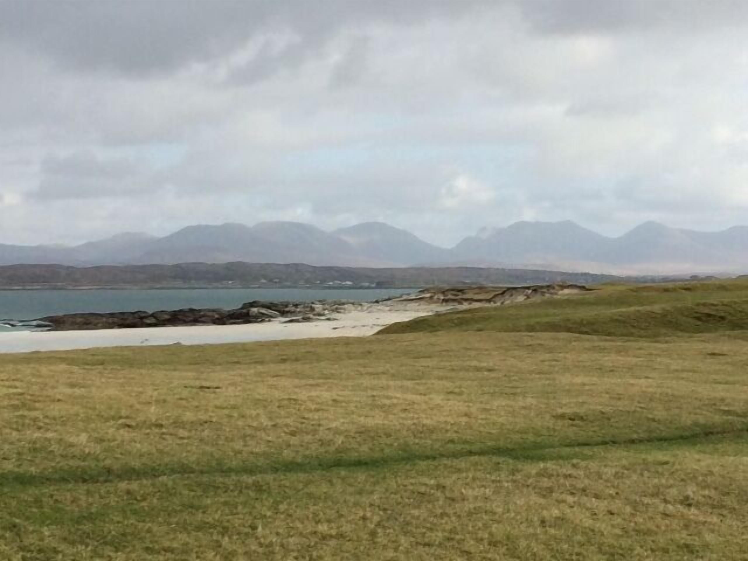A coastal landscape with a grassy foreground, rocky shoreline, and distant mountains under a cloudy sky.