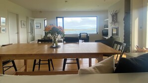 A spacious living room with a large wooden dining table, a comfortable sofa, and a view of the sea through a large window. - Ocean Front Home with Spectacular Views Overlooking Mannin Bay (Ballyconneely)