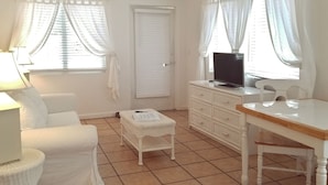 A small, well-lit room with a white sofa, a wicker coffee table, a TV on a stand, a small cabinet, and a window with curtains. - Siesta Key Bungalows: Coquina Bungalows (Sarasota)