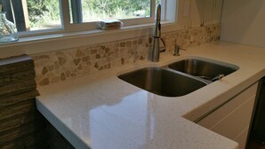Een keuken met een dubbele roestvrijstalen spoelbak, een backsplash van steenmozaïek en een raam met uitzicht op groen.