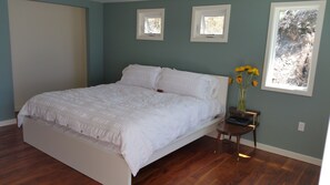 Un dormitorio con cama, mesita de noche, un jarrón con flores y dos ventanas.