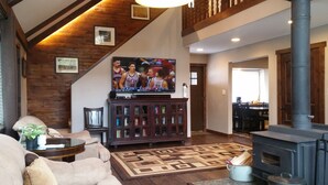 A living room with a fireplace, a television mounted on a wooden cabinet, and a staircase with exposed beams.
