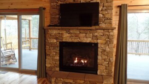 Una chimenea de piedra con un televisor montado encima, suelo de madera y vistas al exterior a través de amplios ventanales.