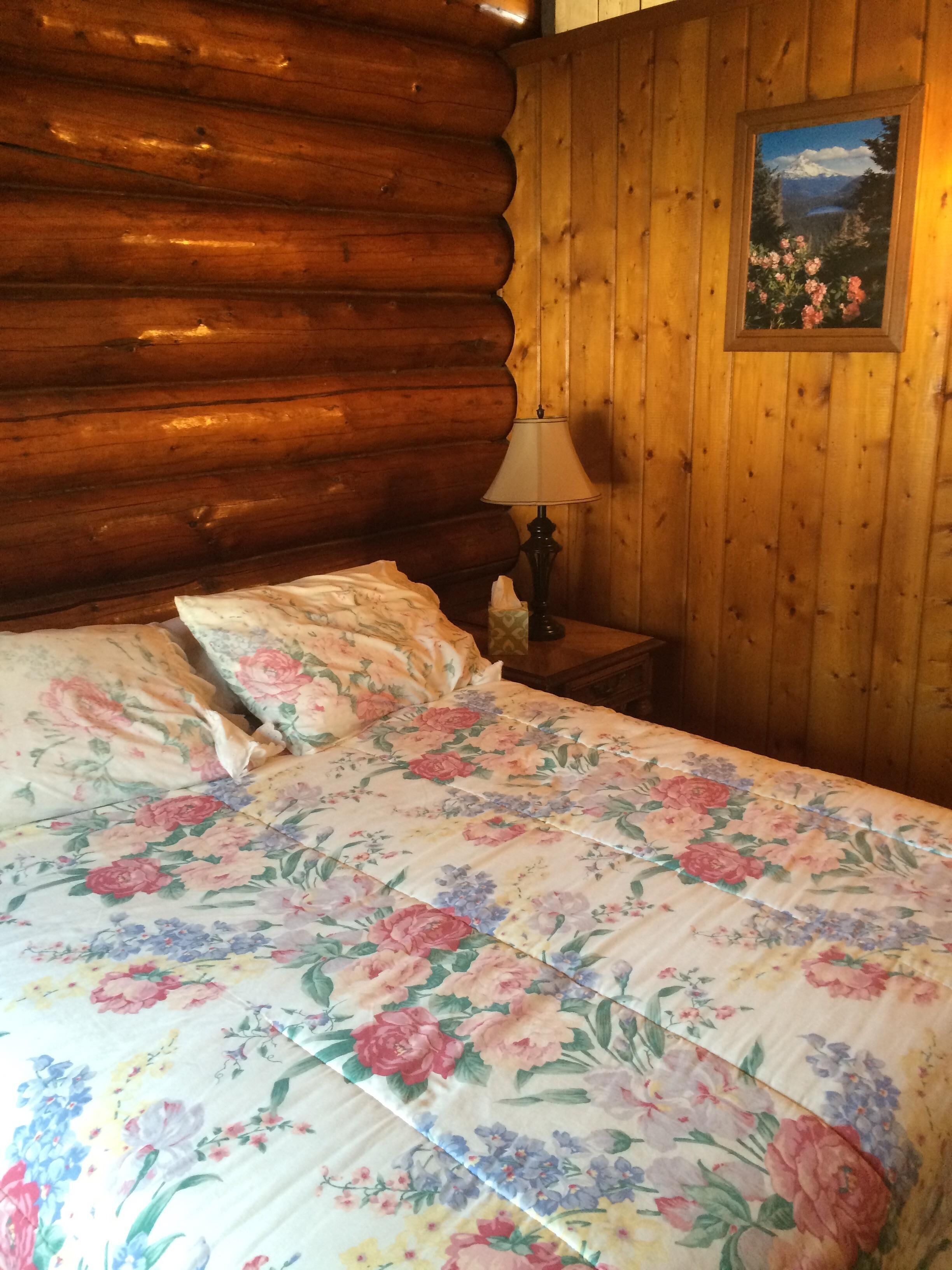 A log cabin room with a bed, a nightstand, a lamp, and a framed picture on the wall.