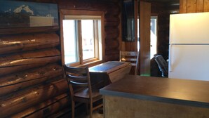 Interno di una baita di legno con un tavolo da pranzo in legno, due sgabelli, un frigorifero e una finestra vista montagne.