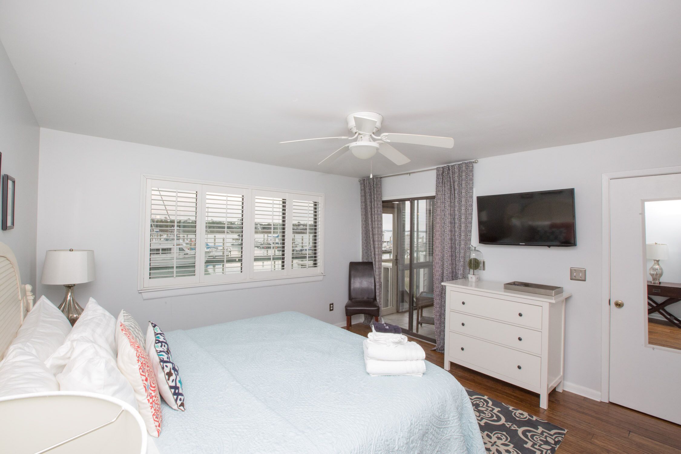 Une chambre  à coucher comprenant un lit, un ventilateur de plafond, une télévision, une chaise, une commode et une fenêtre avec des stores.