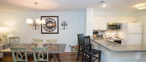 A modern kitchen with white cabinets, a microwave, and a refrigerator. There is a dining area with a table and chairs.