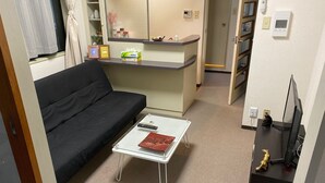 Ein kleines Hotelzimmer mit einer schwarzen Couch, einem kleinen weißen Tisch mit einem Buch, einer Küchenzeile und einem Fernseher auf einem Schrank.
