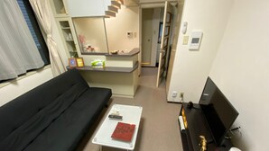 Ein kompakter Wohnbereich mit einem schwarzen Sofa, einem kleinen weißen Couchtisch, einer TV-Konsole mit Fernseher und einer Küchenzeile.