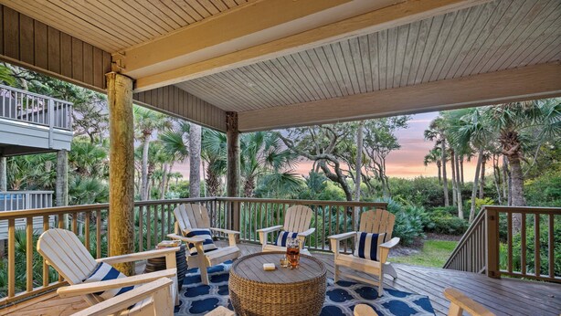 A covered outdoor seating area with Adirondack chairs and a round table, surrounded by lush greenery and a view of palm trees.