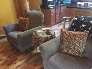 A living room with two grey armchairs, a coffee table, and a leopard print cushion. A kitchen area is visible in the background.