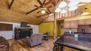 Interior acogedor de una cabaña con chimenea de piedra, vigas de madera en el techo, zona de comedor y cocina con armarios de madera.