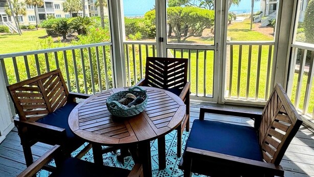 A covered outdoor seating area with a round table and chairs, overlooking a well-maintained lawn and buildings in the background. - Port O' Call D101 by Deserving Vacations (Isle of Palms)