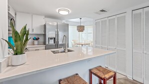 A modern kitchen with white cabinets, a stainless steel refrigerator, and a built-in sink. There is a dining area with a table and chairs, and a potted plant on the counter. - Port O' Call D101 by Deserving Vacations (Isle of Palms)