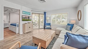 A modern living room with a sofa, a coffee table, and a blue chair. There is a door leading to a bedroom with a bed and a dresser. - Port O' Call D101 by Deserving Vacations (Isle of Palms)
