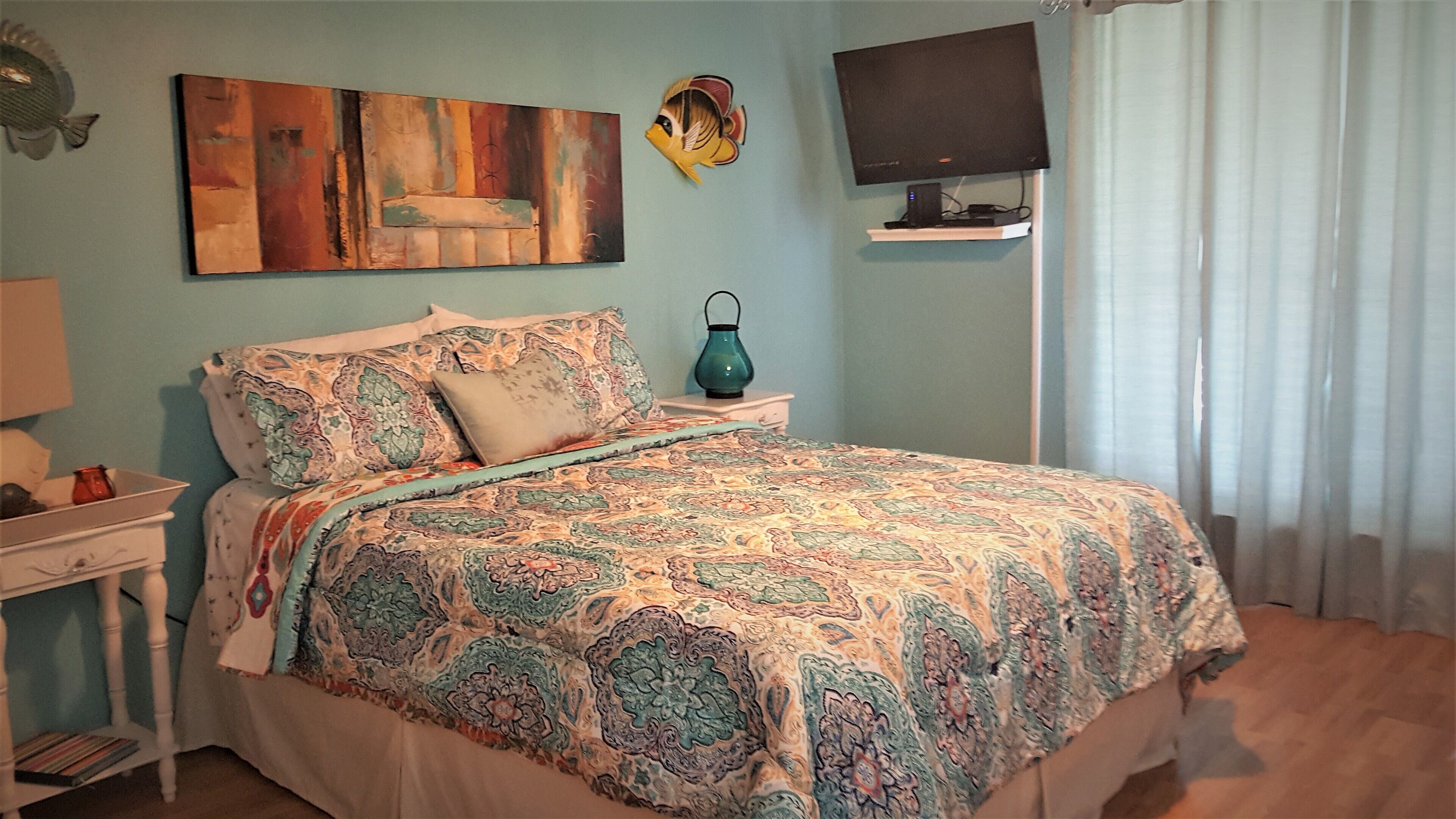 A bedroom with a patterned bedspread, a wall-mounted TV, a fish decoration, and a nightstand with a lamp.