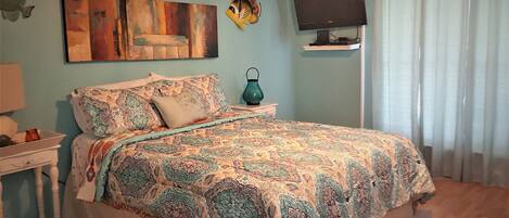 A bedroom with a patterned bedspread, a wall-mounted TV, a fish decoration, and a nightstand with a lamp.