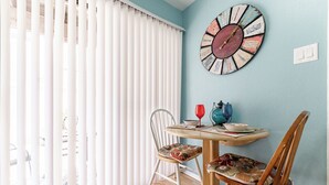 Comedor con mesa redonda, dos sillas, un reloj de pared y una ventana con persianas blancas.