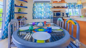 A dining area with a round glass table, chairs with sunflower decorations, and a kitchen in the background.