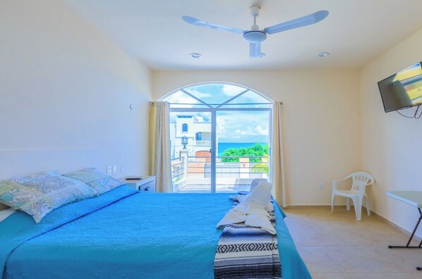 A bedroom with a bed, a ceiling fan, a TV, and a view of the sea through a glass door.
