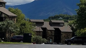A parking lot with a stone building in the center, surrounded by trees and a mountain range in the background.