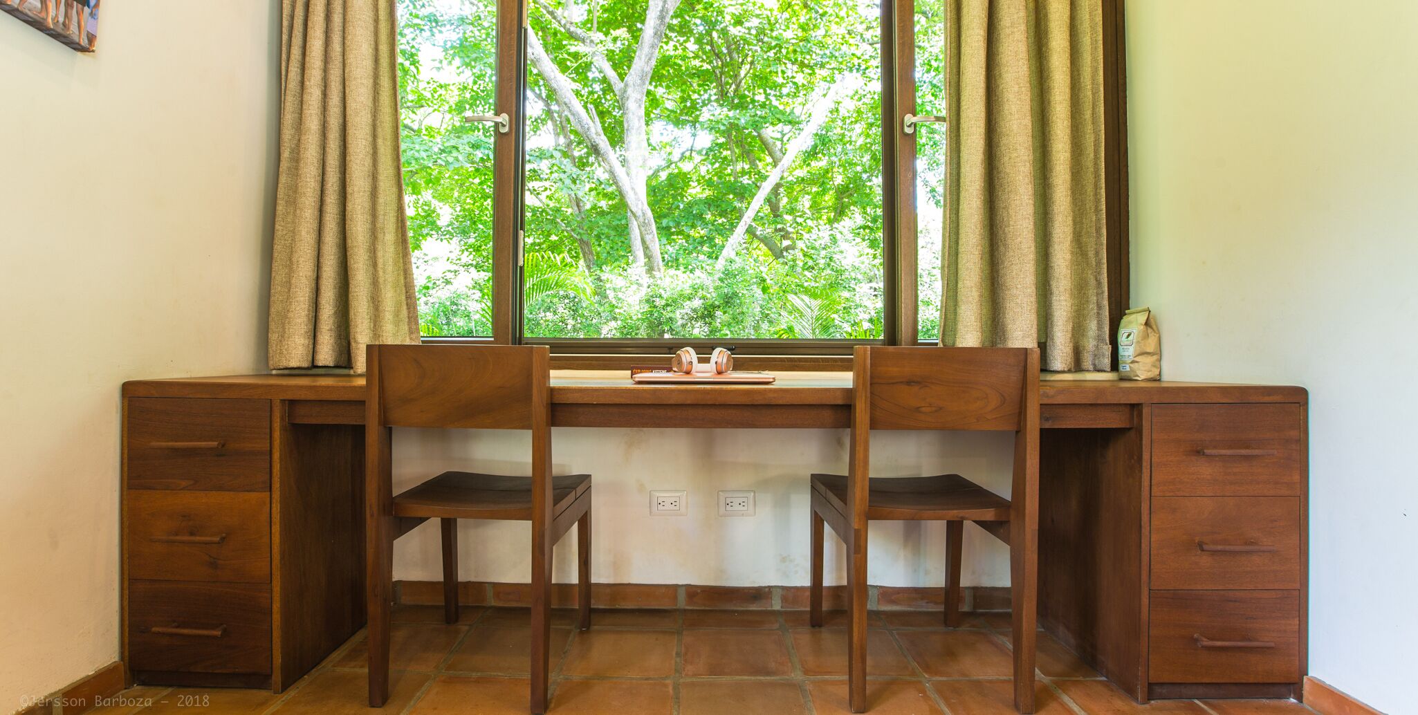 Un escritorio de madera con dos sillas, una ventana con cortinas y vistas a árboles desde afuera.