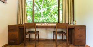 A wooden desk with two chairs, a window with curtains, and a view of trees outside.