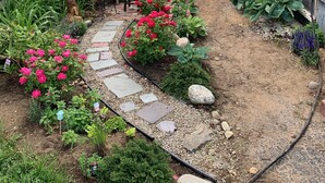 Un sentier de jardin mène à une arche blanche, qui est entourée de différentes plantes et d’une chaise en osier.