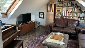 Un salon chaleureux avec un piano, un téléviseur, un canapé en cuir marron, un tapis à motifs et une bibliothèque remplie de livres et d’objets décoratifs.