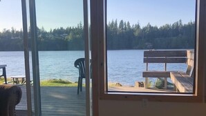Una habitación con un ventanal grande que da a un lago, una mesa de madera con una computadora portátil, una cámara y otros objetos, y una silla junto a la ventana.