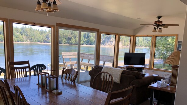 Un comedor con mesa y sillas de madera, un sofá y un televisor. Amplias ventanas ofrecen vistas a un lago y un bosque.