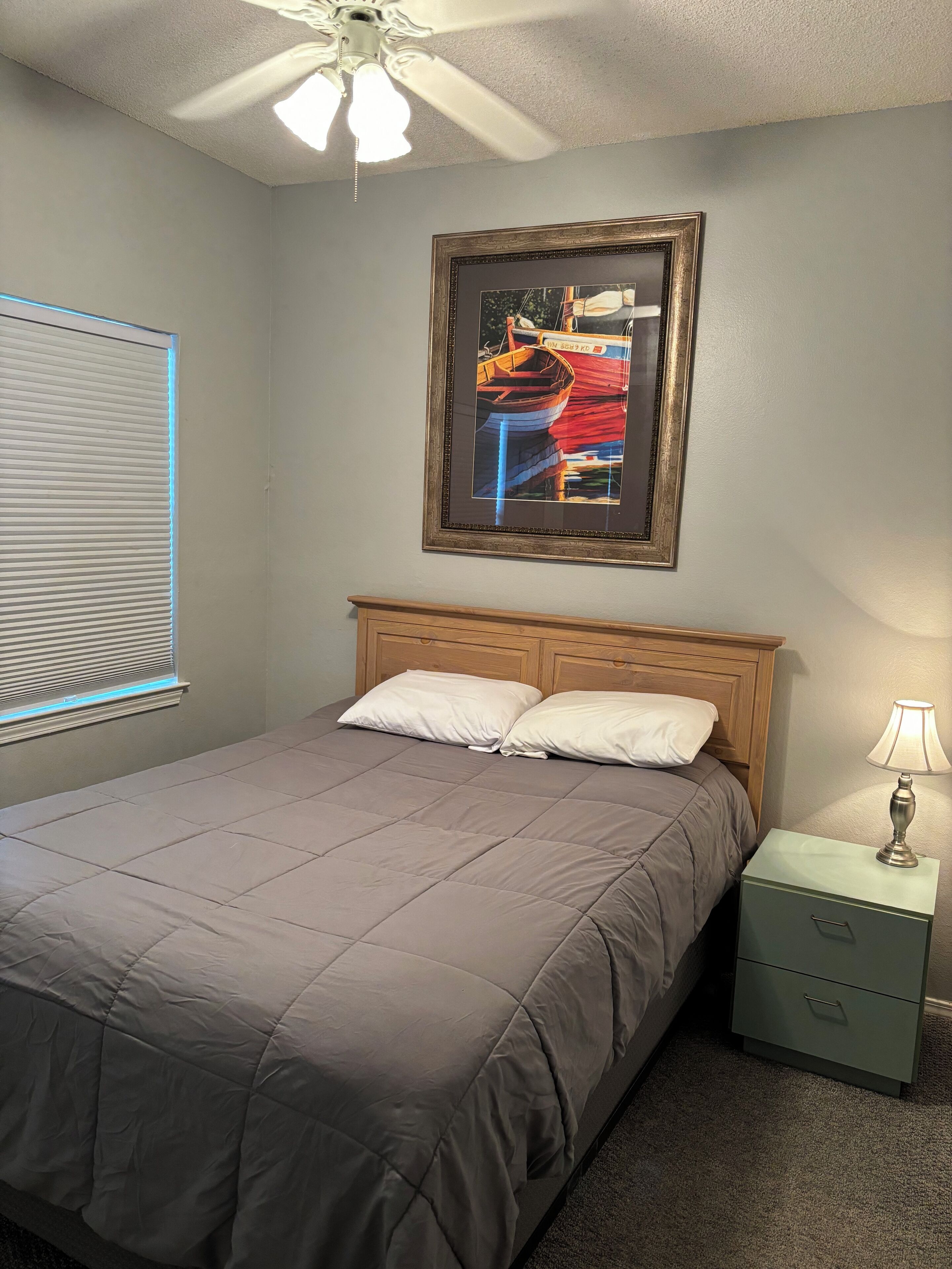 A bedroom with a bed, a nightstand, a lamp, a ceiling fan, and a framed picture on the wall.