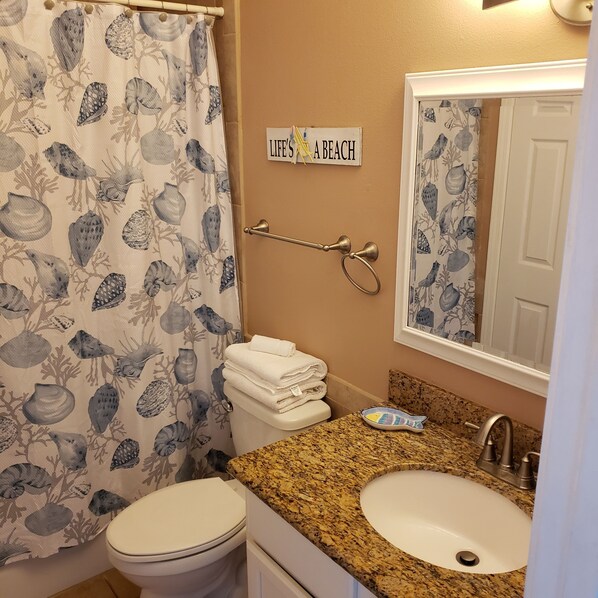 A bathroom with a toilet, sink, and shower curtain with a shell pattern. There's a towel rack with folded towels and a sign that says 'LIFE'S A BEACH'.