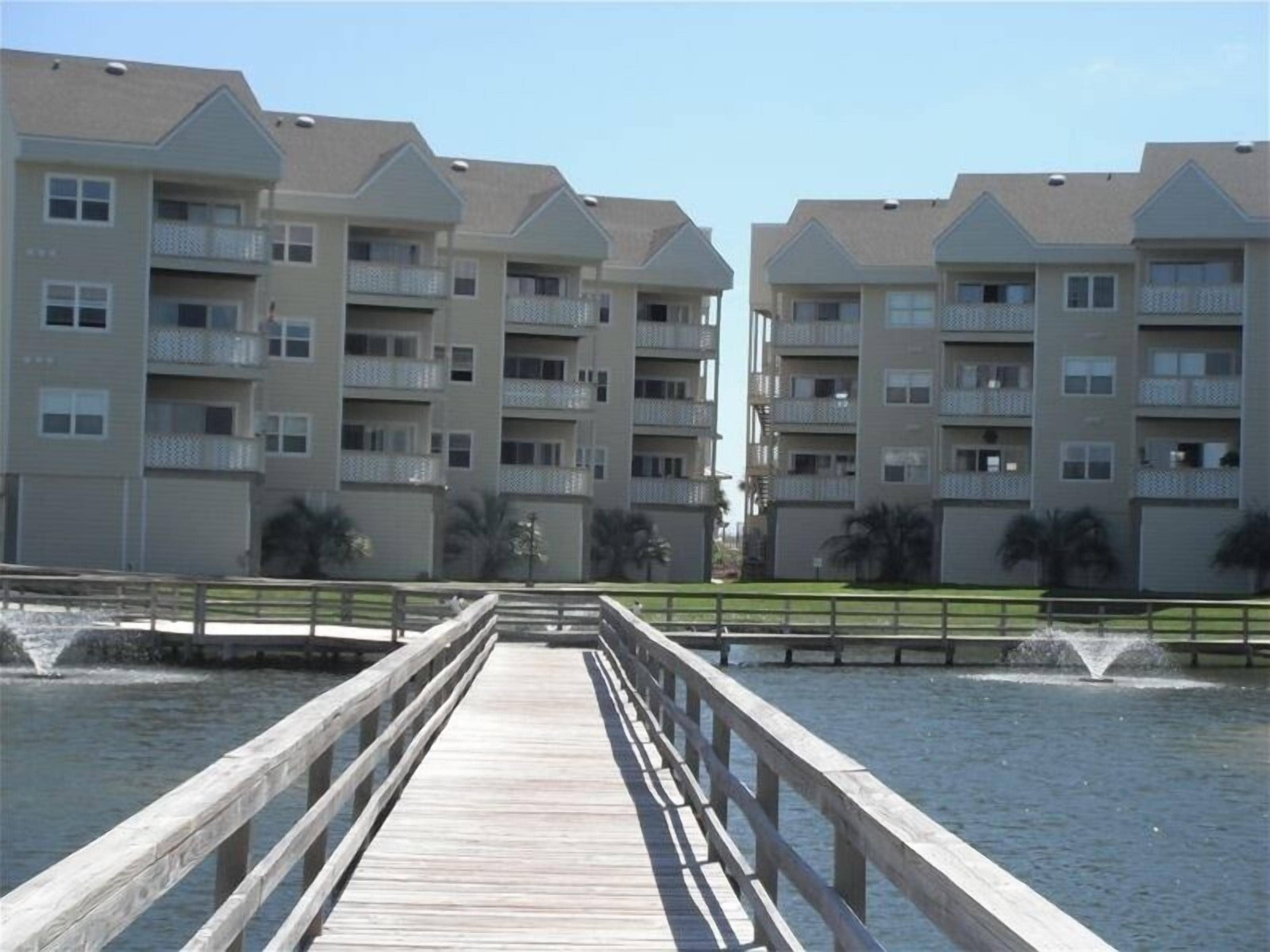 A wooden pier leading to a row of multi-story buildings with balconies, overlooking a body of water.