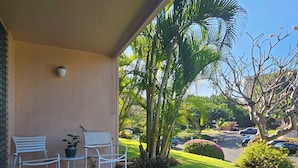A covered patio with two white chairs and a small table, surrounded by lush greenery and palm trees.