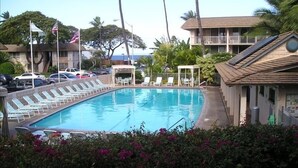 A swimming pool with lounge chairs and a covered seating area, surrounded by palm trees and a building with a balcony.