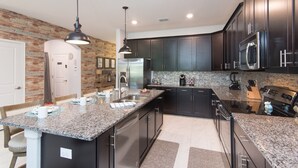 A modern kitchen with dark cabinetry, granite countertops, stainless steel appliances, and a brick accent wall. - Spacious 6BR Family Retreat w/ Private Pool and Spa in Resort Community! (Davenport)