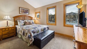 A bedroom with a bed, a wooden nightstand, a bench, a dresser, a lamp, and a large window with a view of mountains. - Spacious Luxury Downtown Riverfront Loft Condo.  License #6027 (Estes Park)