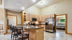 A modern kitchen with wooden cabinets, a central island with bar stools, stainless steel appliances, and a view into the adjacent bedroom. - Spacious Luxury Downtown Riverfront Loft Condo.  License #6027 (Estes Park)