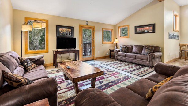 A living room with a brown leather sofa, a wooden coffee table, a TV, and a large window with a view of trees. - Spacious Luxury Downtown Riverfront Loft Condo.  License #6027 (Estes Park)