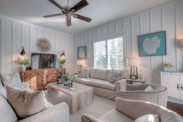 A living room with a sofa, armchairs, a coffee table, and a TV. There is a ceiling fan, a window with blinds, and a wall-mounted TV.