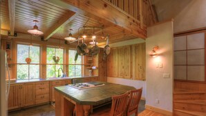 Une cuisine avec des meubles en bois, un grand îlot central doté de chaises, des ustensiles de cuisine suspendus et une vue agréable par les fenêtres.