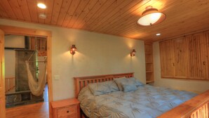 Une chambre aux murs lambrés, avec un lit, deux appliques murales, une table de chevet en bois, un hamac suspendu et une cabine de douche en pierre.